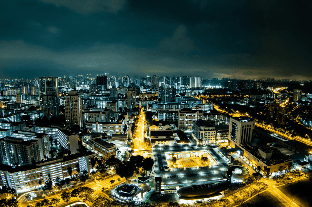 A dazzling nighttime aerial view of Singapore, showcasing the vibrant cityscape near jalan besar, roxy square, and East Coast Road, where iconic laksa stalls like Asia Delight Laksa, Janggut Laksa, Yishun Laksa, Sungei Road Laksa, and Ng Chwee Seng stall opens with unique opening hours. These coffee shop and food centre locations serve Singapore Katong laksa, katong laksa, and Singapore laksa in a regular bowl or small bowl, often with just a spoon or spoon on the side. The laksa recipe features rice noodles, thick bee hoon, and noodles soaked in creamy laksa gravy made from coconut milk, evaporated milk, fresh coconut, and milk, cooked with laksa paste, blue ginger, dried shrimp, dried oysters, sambal, spices, sugar, lard, and garnish, then simmered to perfection. Toppings include fish cake, fishcake, fried tofu puffs, tofu puffs, tau pok, bean sprouts, hard boiled egg, fresh prawns, prawns, cockles, blood cockles, chicken, and seafood. This tasty, savoury, spicy dish is a personal favourite for its flavourful gravy, rich broth, and unforgettable taste—a must-eat food experience in Singapore.