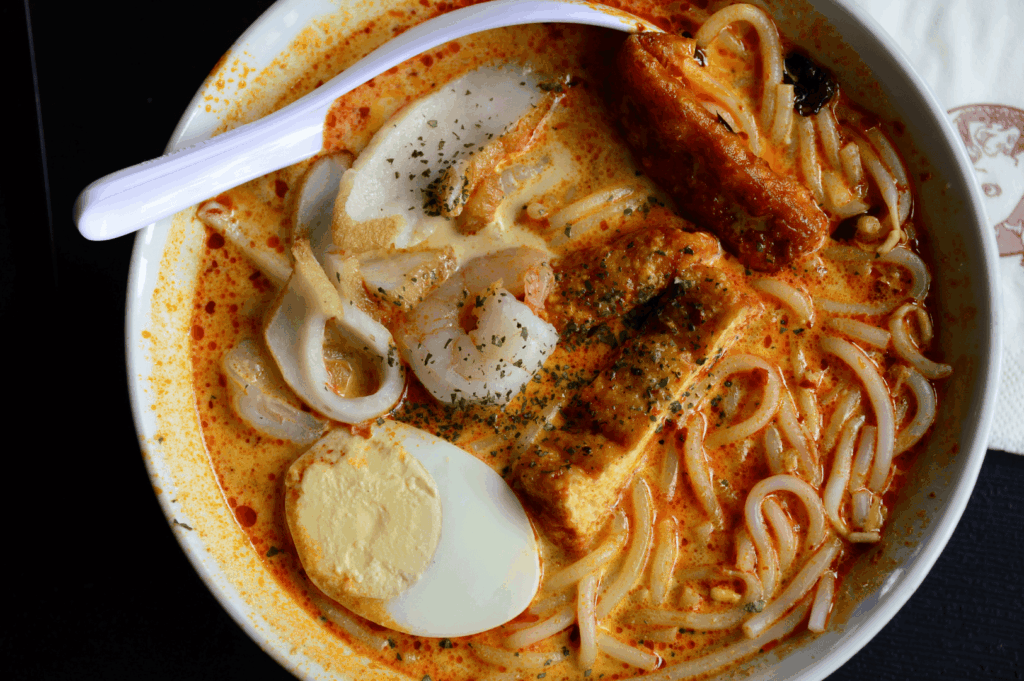 A regular bowl of Singapore Katong laksa served in a coffee shop near East Coast Road, featuring thick rice noodles and thick bee hoon soaked in creamy laksa gravy made with coconut milk, evaporated milk, and fresh coconut. This flavourful, spicy soup is garnished with fried tofu puffs, tofu puffs, tau pok, bean sprouts, hard boiled egg, fish cake, fishcake, fresh prawns, prawns, cockles, blood cockles, and dried shrimp, with hints of dried oysters, blue ginger, and laksa paste. A spoon rests on the side—just a spoon—ready to eat this savoury, tasty dish. Popular at laksa stalls like Janggut Laksa, Asia Delight Laksa, Yishun Laksa, Sungei Road Laksa, and Ng Chwee Seng, this laksa recipe is a personal favourite among seafood lovers in Singapore. Found in food centres, jalan besar, and roxy square, each stall opens with unique opening hours, offering lemak gravy, cooked with spices, chicken, otah, lard, milk, sugar, and garnish, then simmered to perfection. The flavour, taste, and curry depth make this Singapore laksa a must-try.