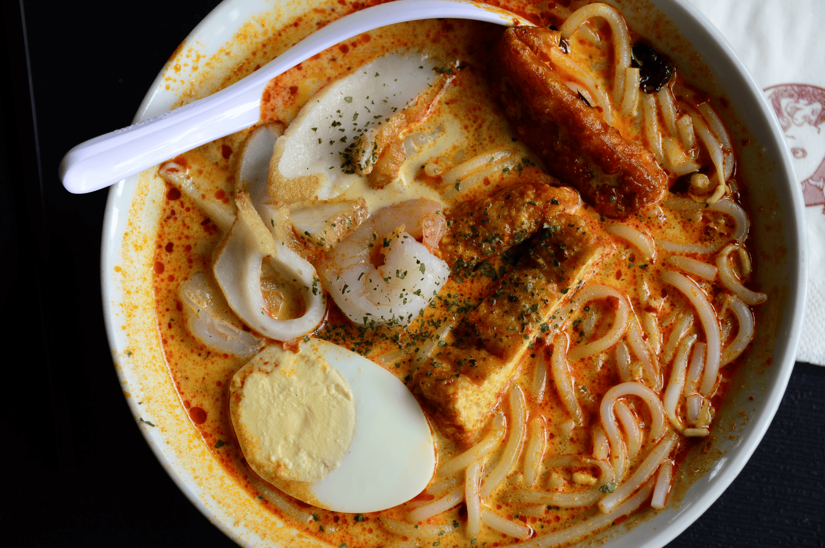 A regular bowl of Singapore Katong laksa served in a coffee shop near East Coast Road, featuring thick rice noodles and thick bee hoon soaked in creamy laksa gravy made with coconut milk, evaporated milk, and fresh coconut. This flavourful, spicy soup is garnished with fried tofu puffs, tofu puffs, tau pok, bean sprouts, hard boiled egg, fish cake, fishcake, fresh prawns, prawns, cockles, blood cockles, and dried shrimp, with hints of dried oysters, blue ginger, and laksa paste. A spoon rests on the side—just a spoon—ready to eat this savoury, tasty dish. Popular at laksa stalls like Janggut Laksa, Asia Delight Laksa, Yishun Laksa, Sungei Road Laksa, and Ng Chwee Seng, this laksa recipe is a personal favourite among seafood lovers in Singapore. Found in food centres, jalan besar, and roxy square, each stall opens with unique opening hours, offering lemak gravy, cooked with spices, chicken, otah, lard, milk, sugar, and garnish, then simmered to perfection. The flavour, taste, and curry depth make this Singapore laksa a must-try.
