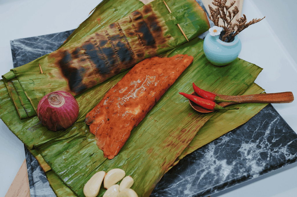 A vibrant otah on a banana leaf, surrounded by blue ginger, red chili, garlic, and onion—ingredients often used in laksa recipes across Singapore. This dish, commonly found at laksa stalls like Asia Delight Laksa, Janggut Laksa, Yishun Laksa, Sungei Road Laksa, and Ng Chwee Seng, is cooked with laksa paste, coconut milk, evaporated milk, fresh coconut, and milk, then simmered into creamy laksa gravy. Served in a regular bowl or small bowl with just a spoon or spoon, Singapore Katong laksa and katong laksa feature rice noodles, thick bee hoon, and noodles soaked in spicy, flavourful broth. Toppings include fish cake, fishcake, fried tofu puffs, tofu puffs, tau pok, bean sprouts, hard boiled egg, fresh prawns, prawns, cockles, blood cockles, chicken, and seafood, garnished with sambal, dried shrimp, dried oysters, sugar, lard, and other spices. Found in coffee shop and food centre settings near east coast road, jalan besar, and roxy square, each stall opens with unique opening hours, offering a tasty, savoury personal favourite that’s a must-eat in Singapore for its unforgettable taste, curry depth, and lemak richness.
