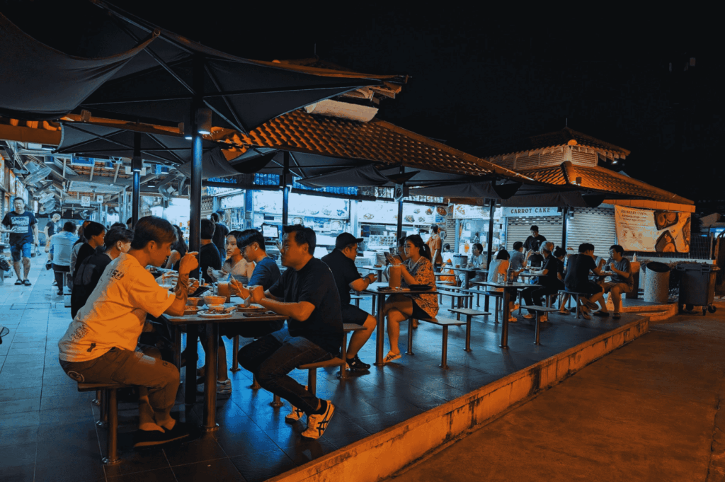 Bustling open air food court at night in one of Singapore's iconic hawker centres, featuring street food vendors and hawker stalls serving char kway teow with chinese sausage, hainanese chicken rice, nasi lemak with coconut milk, and other local dishes. Diners enjoy mouthwatering food like stir fried flat rice noodles with dark soy sauce, chili sauce, and bean sprouts, surrounded by illuminated food stalls in a vibrant food centre near the central business district. This real life scene captures the essence of hawker culture at places like lau pa sat, maxwell food centre, and airport road food centre—must visit spots for great food, drinks like teh tarik and coffee, and dishes such as roti prata, hokkien mee, chilli crab, and shaved ice. A short walk from orchard road or the singapore river, these hawker centers offer delicious dinner options, hidden gems, and local cuisine celebrated by anthony bourdain and travelers across southeast asia. Ideal for blog post travel tips on singapore's hawker centers and public transport access to surrounding islands and chinatown.