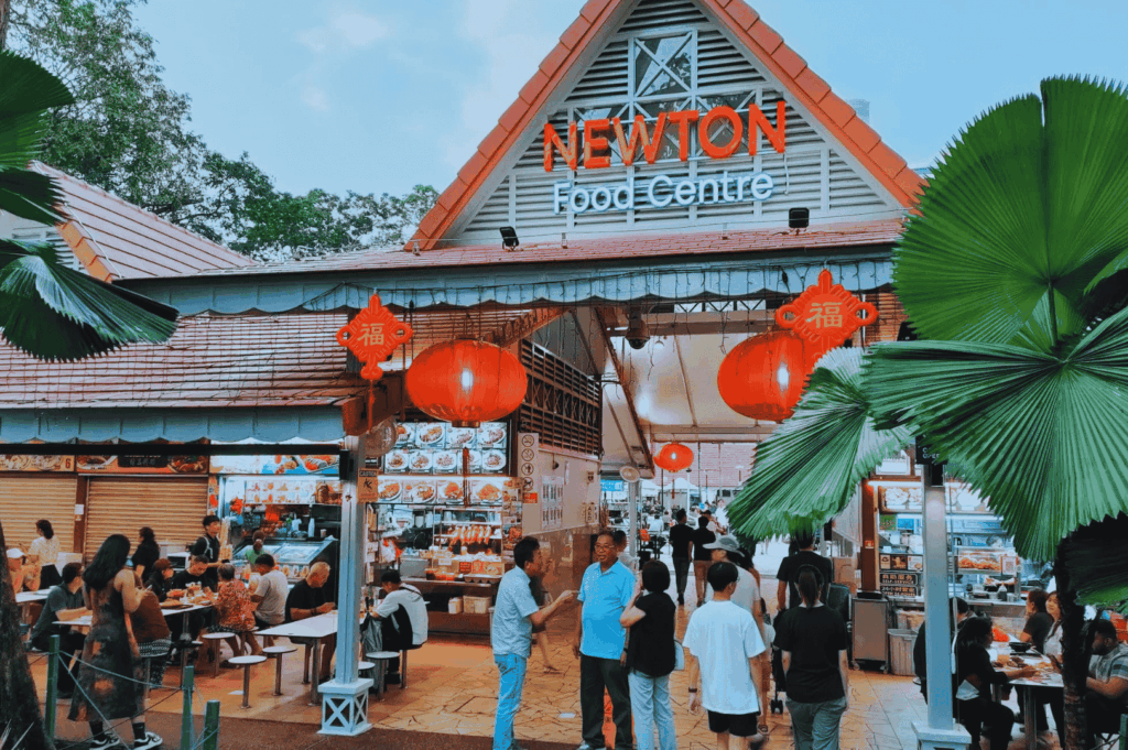 Entrance of Newton Food Centre, one of Singapore's iconic hawker centres, bustling with street food vendors and diners enjoying hawker food like char kway teow with chinese sausage, hainanese chicken rice, nasi lemak with coconut milk, and other local dishes. Red lanterns and festive decor highlight the vibrant hawker culture and open air food courts that define singapore's hawker centers. Food stalls inside serve stir fried flat rice noodles with dark soy sauce, chili sauce, and bean sprouts, alongside malay dishes, roti prata, chilli crab, hokkien mee, and shaved ice. Located a short walk from orchard road and the central business district, Newton is a must visit spot for mouthwatering food and drinks like teh tarik and coffee. This food centre, like lau pa sat, maxwell food centre, and airport road food centre, offers great food, hidden gems, and real life dining experiences praised by anthony bourdain. Perfect for travel tips and blog post guides on public transport access to surrounding islands, the singapore river, little india, and chinatown. Most stalls serve delicious noodles, rice, shrimp, egg, and chicken dishes with soy sauce and other ingredients from southeast asia and the world.