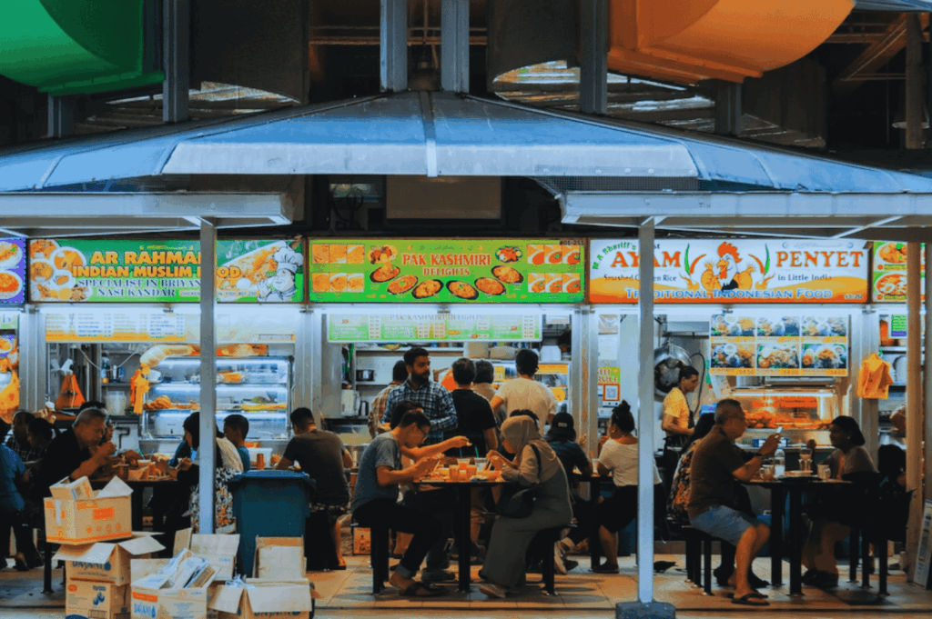 Indoor food centre in Singapore filled with street food vendors at brightly lit hawker stalls serving mouthwatering food like nasi lemak with coconut milk, hainanese chicken rice, ayam penyet, and malay dishes; stalls such as AR RAHMAN INDIAN MUSLIM FOOD and PAK KASHMIRI BRIYANI showcase local cuisine and other dishes served with rice, egg, and chili sauce; diners enjoy hawker food at communal tables in one of singapore's iconic hawker centres, reflecting the vibrant hawker culture found at airport road food centre, maxwell food centre, and lau pa sat near the singapore river and central business district; a real life scene of singapore's hawker centers offering great food, hidden gems, and local dishes like char kway teow with chinese sausage, flat rice noodles stir fried in dark soy sauce and soy sauce, alongside roti prata, hokkien mee, chilli crab, and shaved ice—perfect for travel tips, blog post inspiration, and anthony bourdain-style food adventures across southeast asia and surrounding islands.