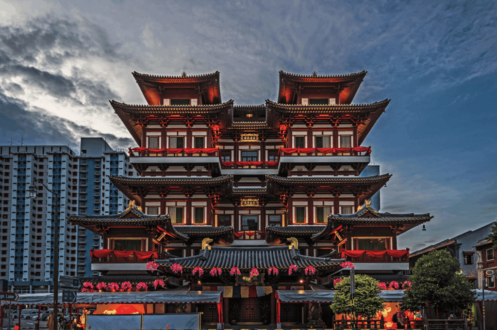 Buddha Tooth Relic Temple in Singapore’s Chinatown, glowing with warm red-and-gold lighting and adorned with lanterns and floral decorations—its ornate Tang-style tiers rise against a backdrop of modern skyscrapers and dramatic skies, symbolizing the city’s reverent blend of spiritual heritage and contemporary skyline.