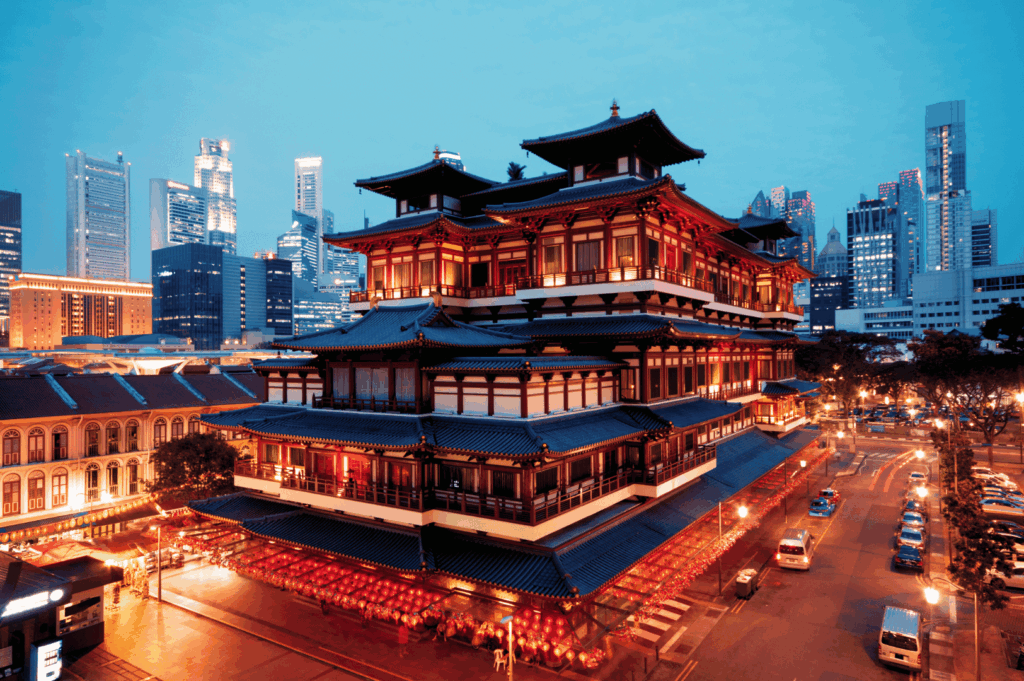 Night view of the Buddha Tooth Relic Temple in Singapore’s Chinatown, glowing with warm red-and-gold illumination and surrounded by festive lanterns—set against a backdrop of sleek skyscrapers, this scene captures the city’s striking contrast between sacred heritage and modern urban life.