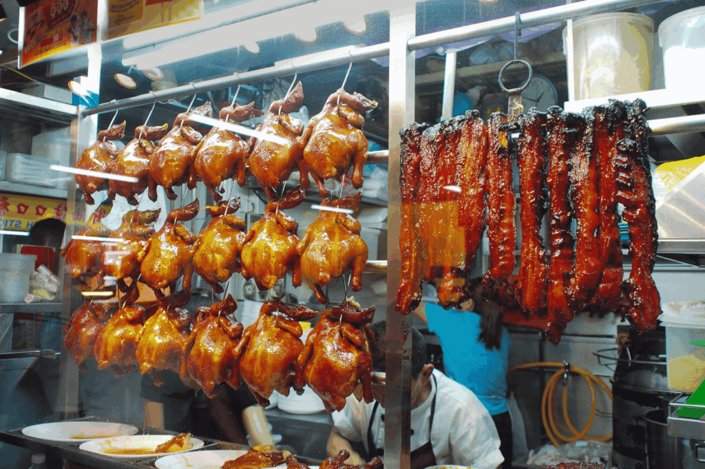Display of whole roasted ducks and char siu pork hanging behind glass at a Chinatown hawker stall in Singapore—golden-brown glazes glisten under market lights as cooks prepare food in the background, capturing the rich culinary traditions that thrive amid the city’s modern bustle