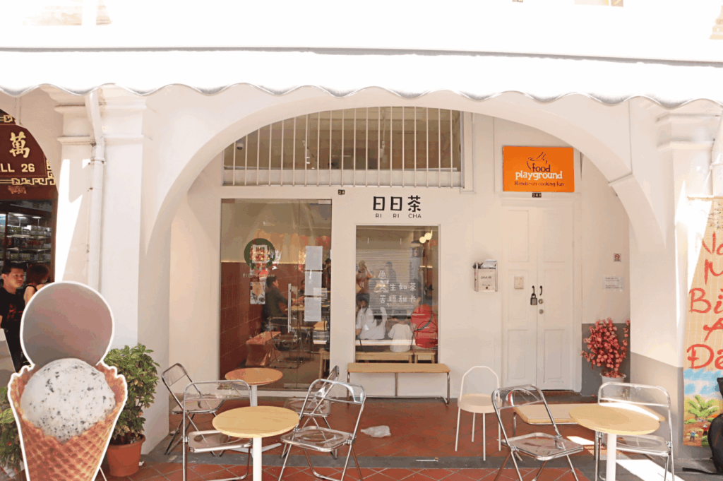 Exterior of Ri Ri Cha tea shop in Singapore’s Chinatown, with arched white façade, outdoor seating, and a glimpse into its cozy interior—this modern café blends minimalist design with local character, reflecting the district’s evolving food scene and everyday cultural rhythms.