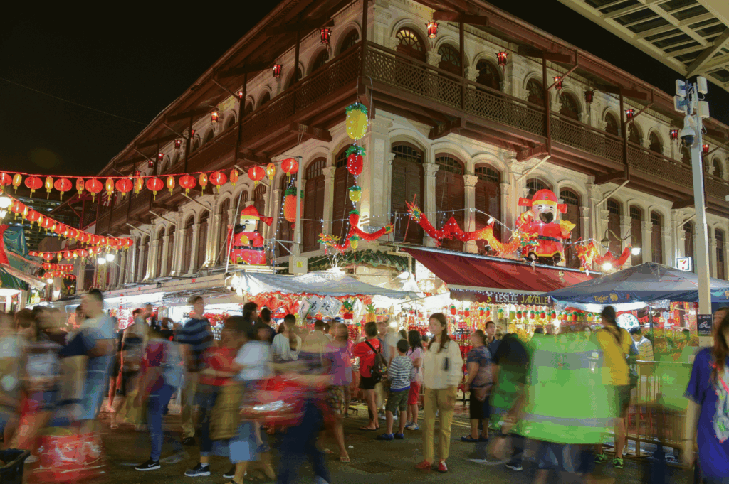 Lunar New Year night market in Singapore’s Chinatown, bustling with crowds beneath red lanterns and colorful lights—historic colonial buildings glow warmly behind inflatable Chinese deities and festive stalls, capturing the district’s joyful fusion of heritage celebration and urban vibrancy.