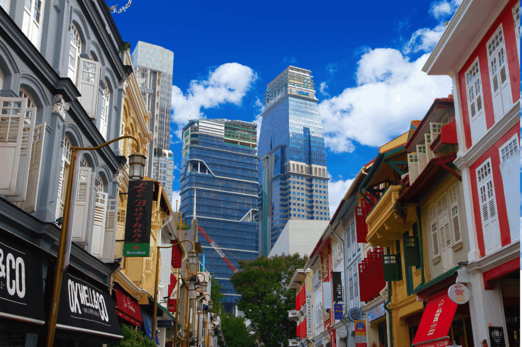 Colorful colonial shophouses line a lively Singapore street, their ornate facades and heritage signage juxtaposed against towering glass skyscrapers—capturing the layered coexistence of tradition and innovation in Chinatown’s evolving urban landscape.