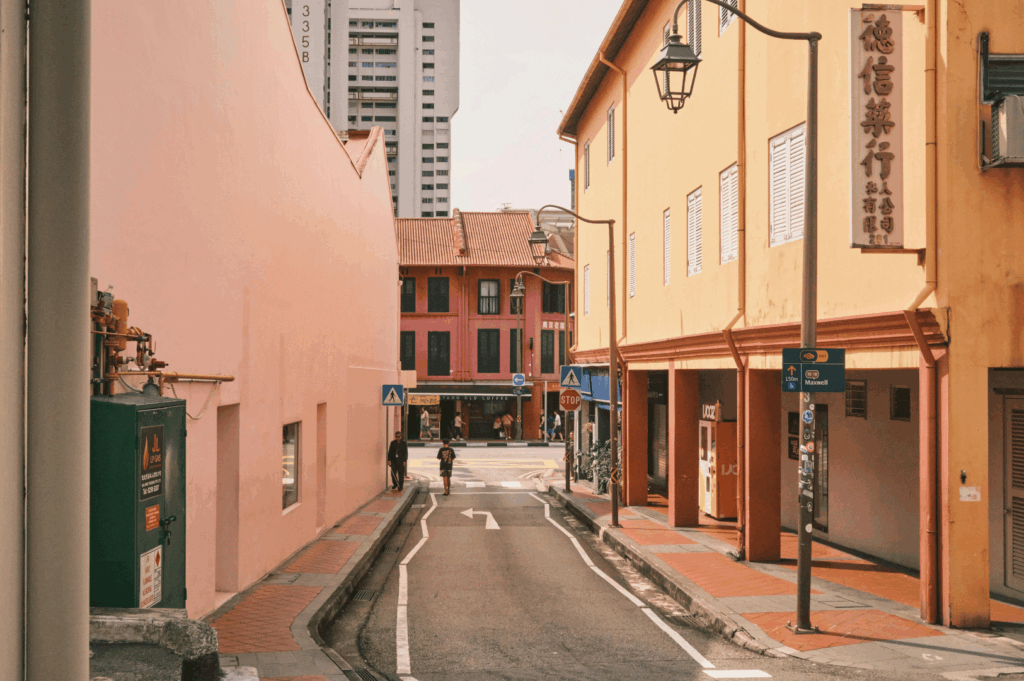 Narrow Chinatown street in Singapore lined with warm-toned heritage buildings and Chinese signage, where pedestrians and cyclists move through a quiet urban scene—framed by traditional facades in the foreground and a modern high-rise in the distance, reflecting the district’s layered architectural identity.