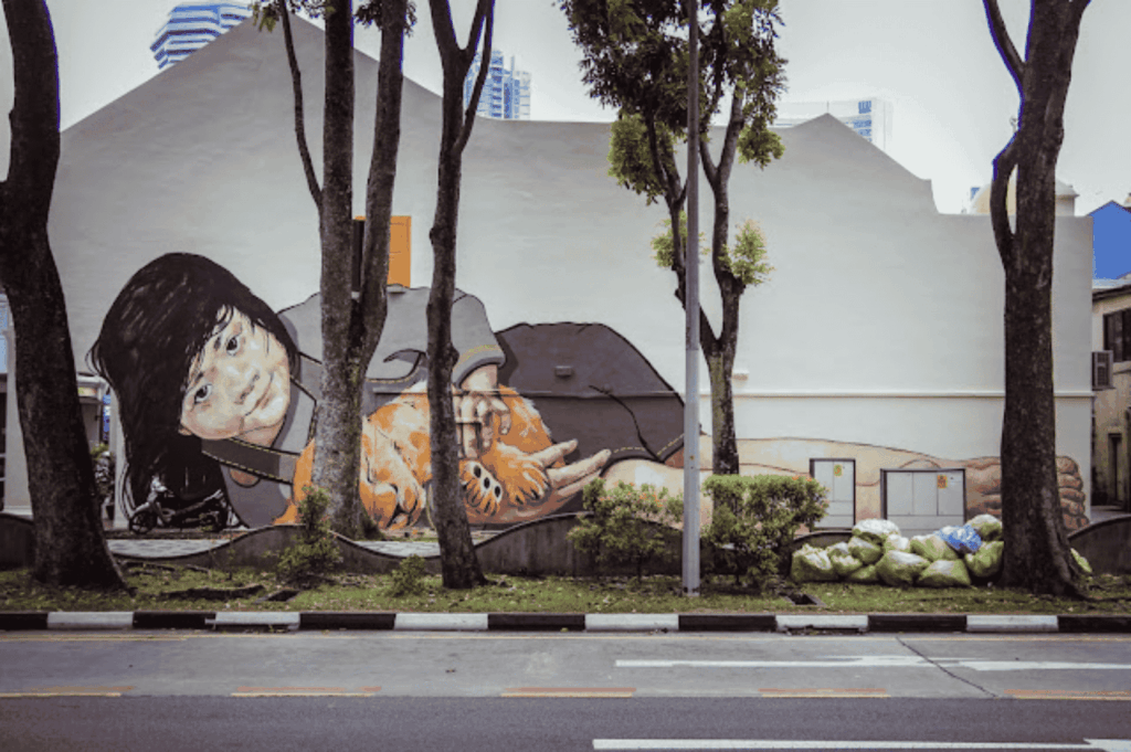 Massive mural painted on the wall of a modern city building, depicting a realistic figure lying down holding an orange cat, blending emotional storytelling with the scale of urban art in the Singapore art scene. Surrounded by public spaces, streets, and main road views, this striking work reflects the diversity of art forms in art in Singapore—from street art, graffiti, and singapore street art on Victoria Street, Little India, Haji Lane, Clive Street, Kerbau Road, Rowell Road, Ann Siang Hill, Tiong Bahru, and Chinatown, to fine arts and performing arts at the Singapore Art Museum, National Gallery Singapore, and during the Singapore Biennale. Created by local and international artists—including Yip Yew Chong, Ernest Zacharevic, and Novena Angela—who have helped develop the contemporary scene in a modern way, it celebrates the multicultural society of Singapore, a young nation in the Asia Pacific region and Southeast Asia. Influences from Chinese immigrants, India, China, and Asia merge traditional and art deco ideas with modern design, supported by the National Heritage Board, Nanyang Academy, and Art SG. The work connects history, culture, life, music, food, and sense of place, playing an important role in linking early exhibitions to today’s collection of sculptures, shops, boutique shops, local neighbourhood events, and global world art moments—even inspiring pop culture like Crazy Rich Asians.