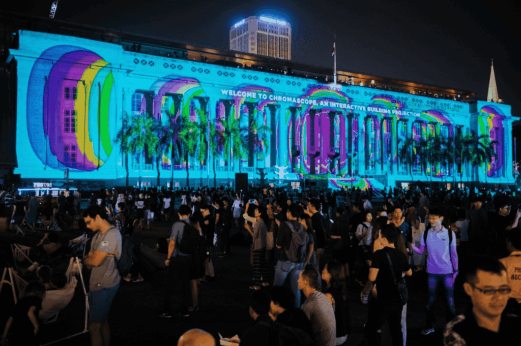 Nighttime public spaces event in the central business district of Singapore, where a historic building is illuminated with vibrant, abstract light projections during an art festival, showcasing the diversity of the Singapore art scene. This large‑scale urban art display blends technology with art forms from fine arts to performing arts, much like works at the Singapore Art Museum, National Gallery Singapore, and during the Singapore Biennale. Surrounded by palm‑lined streets, main road views, and boutique shops, the scene reflects the multicultural society of a young nation in the Asia Pacific region and Southeast Asia, influenced by Chinese immigrants, India, China, and Asia. Similar to street art, graffiti, and singapore street art found on Victoria Street, Little India, Haji Lane, Clive Street, Kerbau Road, Rowell Road, Ann Siang Hill, Tiong Bahru, and Chinatown, the projection celebrates local and international artists—including Yip Yew Chong, Ernest Zacharevic, and Novena Angela—who have helped develop art in Singapore in a modern way. Supported by the National Heritage Board, Nanyang Academy, and Art SG, the event highlights emerging artists, the new generation, and international artists through art exhibitions, collection showcases, sculptures, mural and painted wall works, music, food, and life. Drawing on traditional and art deco ideas, it plays an important role in connecting early exhibitions to today’s contemporary scene, inspired by history, culture, shops, local neighbourhood events, and even pop culture moments like Crazy Rich Asians, cementing Singapore’s place in the world of creativity.