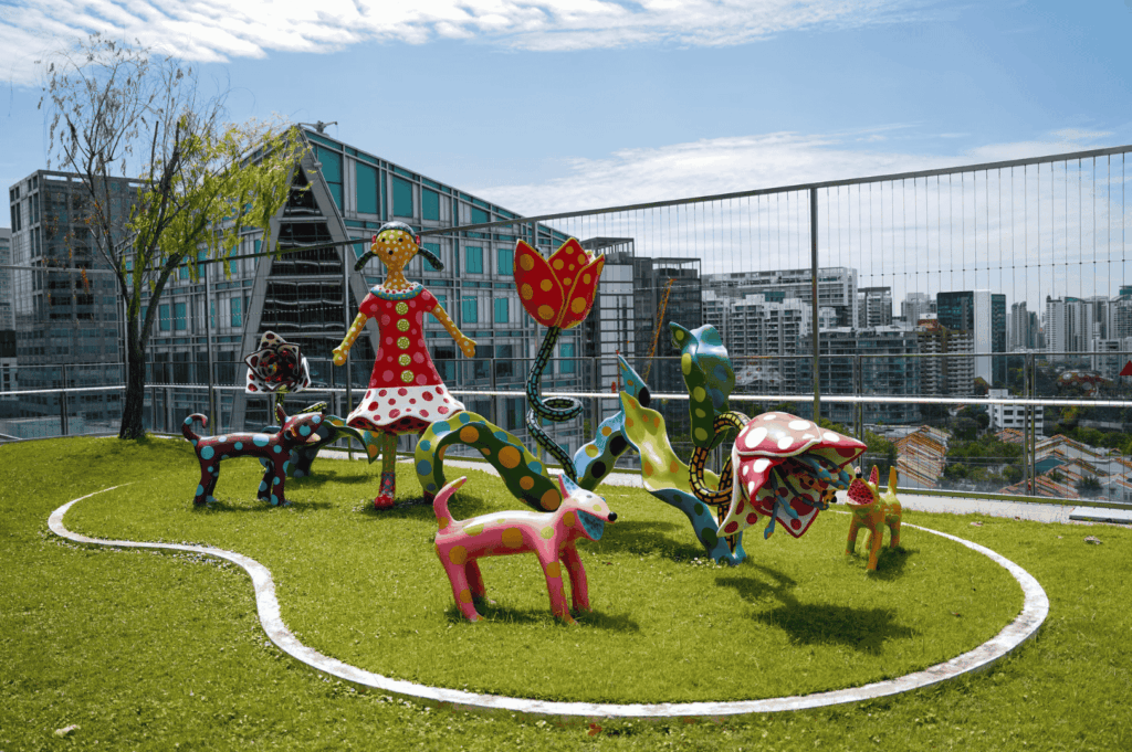 Colorful rooftop public spaces installation in the modern city of Singapore, featuring playful sculptures of a woman in a red polka‑dot dress, stylized animals, oversized flowers, and mushrooms, set against an urban skyline. Part of the vibrant Singapore art scene, this imaginative display blends art forms from fine arts to performing arts, much like works at the Singapore Art Museum, National Gallery Singapore, and during the Singapore Biennale. Surrounded by streets, shops, and boutique shops in neighbourhood hubs such as Victoria Street, Little India, Haji Lane, Clive Street, Kerbau Road, Rowell Road, Ann Siang Hill, Tiong Bahru, and Chinatown, it reflects the multicultural society of a young nation in the Asia Pacific region and Southeast Asia. Influences from Chinese immigrants, India, China, and Asia merge traditional and art deco ideas with a modern way of storytelling, similar to street art, urban art, graffiti, and singapore street art by local and international artists—including Yip Yew Chong, Ernest Zacharevic, and Novena Angela—who have helped develop art in Singapore. Supported by the National Heritage Board, Nanyang Academy, and Art SG, the installation celebrates emerging artists, the new generation, and international artists through art exhibitions, collection highlights, mural and painted wall works, music, food, and life. From early exhibitions to today’s contemporary scene, this inspired work plays an important role in connecting history, culture, main street and main road corridors, local events, and even pop culture moments like Crazy Rich Asians, cementing Singapore’s place in the world of creativity.