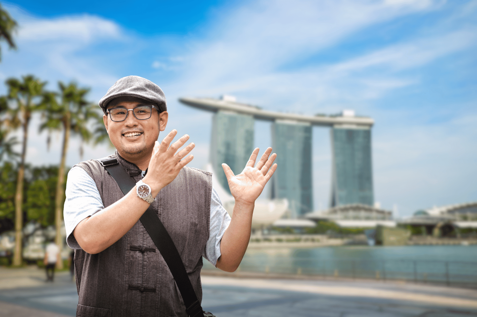 A smiling man stands in front of Marina Bay Sands, gesturing with both hands during a sunny day in Singapore. Dressed casually with a gray cap, glasses, and vest, he embodies the spirit of local exploration. Behind him, the iconic skyline and palm-lined promenade evoke the vibrant energy of Singapore’s evolving community heritage walks.