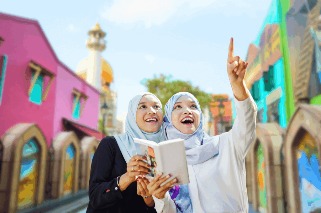 Two women wearing hijabs stand in a vibrant heritage district, holding a book together as one points upward with enthusiasm. Their expressions reflect curiosity and connection, framed by colorful architecture and a golden-domed landmark that hints at Singapore’s rich cultural tapestry. The scene captures the spirit of learning and community engagement central to Singapore’s growing heritage walk movement.