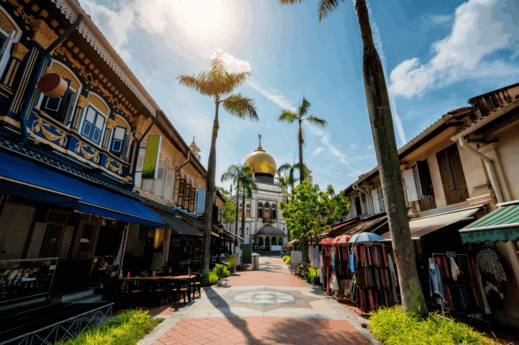 A sunlit pedestrian street in Kampong Glam leads to the majestic Sultan Mosque, framed by tall palm trees and vibrant shophouses with colorful facades and textile displays. The golden dome gleams against a clear sky, anchoring the scene in Islamic architectural heritage. The lively atmosphere reflects Singapore’s fusion of tradition and modernity, capturing the essence of its community heritage walks.