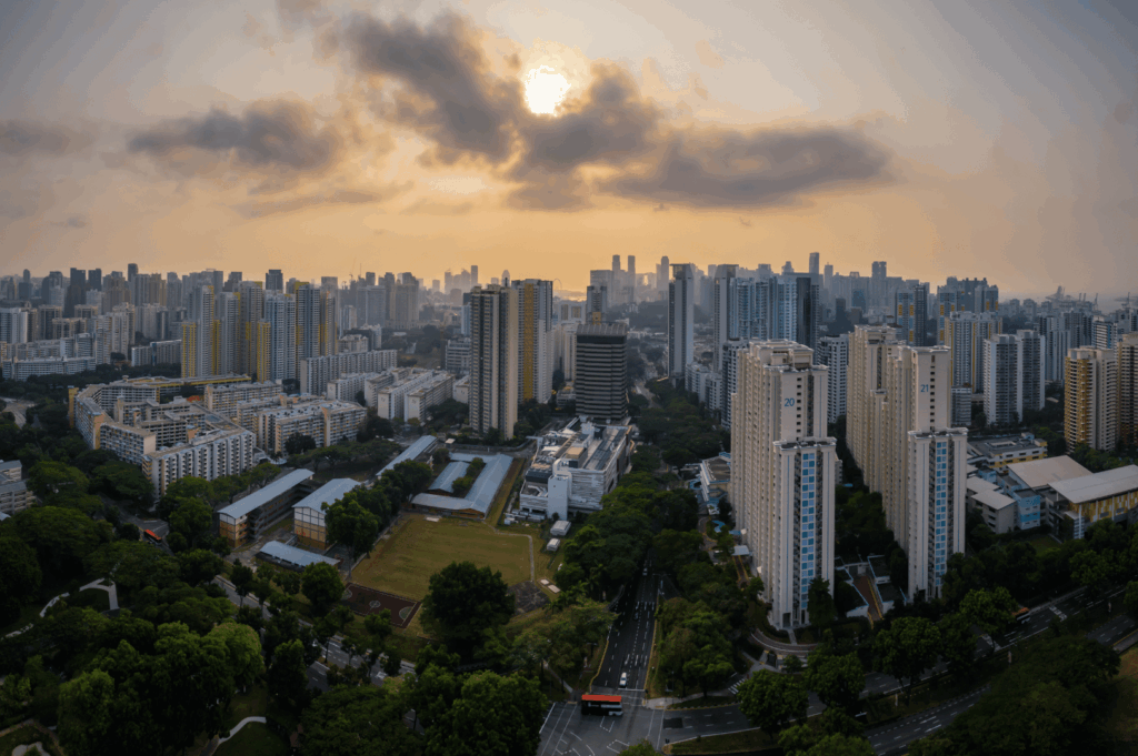 Panoramic view of Singapore’s urban skyline at golden hour, with high-rise buildings bathed in warm sunlight and clouds diffusing the glow. In the foreground, a sports field and tree-lined streets with a red bus hint at everyday life amid dense development. The image captures the interplay between nature and city planning, reflecting the layered textures of community heritage walks in a modern metropolis.