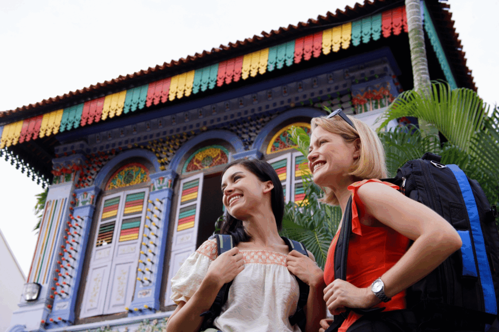 Two travelers stand before a vividly colored heritage building adorned with arched windows, ornate shutters, and a festive roof fringe in red, yellow, and green. Their casual attire and backpacks suggest discovery and curiosity, while lush greenery adds a tropical warmth. The scene celebrates Singapore’s layered cultural identity and the joyful spirit of community heritage walks.
