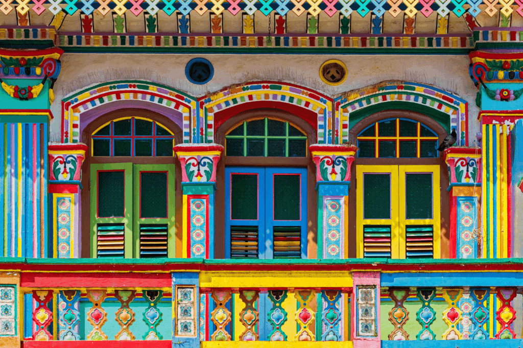 Vibrant facade of a heritage shophouse in Singapore, featuring three arched windows framed by multicolored geometric patterns and a decorative balcony with intricate latticework. The vivid palette—greens, reds, yellows, and blues—reflects the multicultural spirit of the neighborhood, evoking Peranakan, Indian, and Chinese design influences. A visual celebration of Singapore’s layered cultural identity.