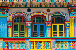 Vibrant facade of a heritage shophouse in Singapore, featuring three arched windows framed by multicolored geometric patterns and a decorative balcony with intricate latticework. The vivid palette—greens, reds, yellows, and blues—reflects the multicultural spirit of the neighborhood, evoking Peranakan, Indian, and Chinese design influences. A visual celebration of Singapore’s layered cultural identity.