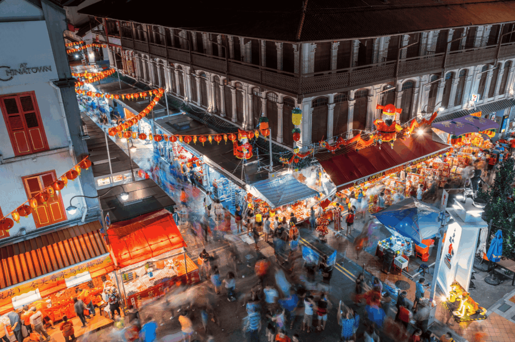 Nighttime street market in Singapore’s Chinatown, glowing with red and yellow lanterns strung overhead. Crowds weave through brightly lit stalls selling food and goods, framed by heritage buildings with traditional balconies and ornate facades. The scene pulses with energy and cultural depth, capturing the communal spirit and festive ambiance of Singapore’s living heritage.
