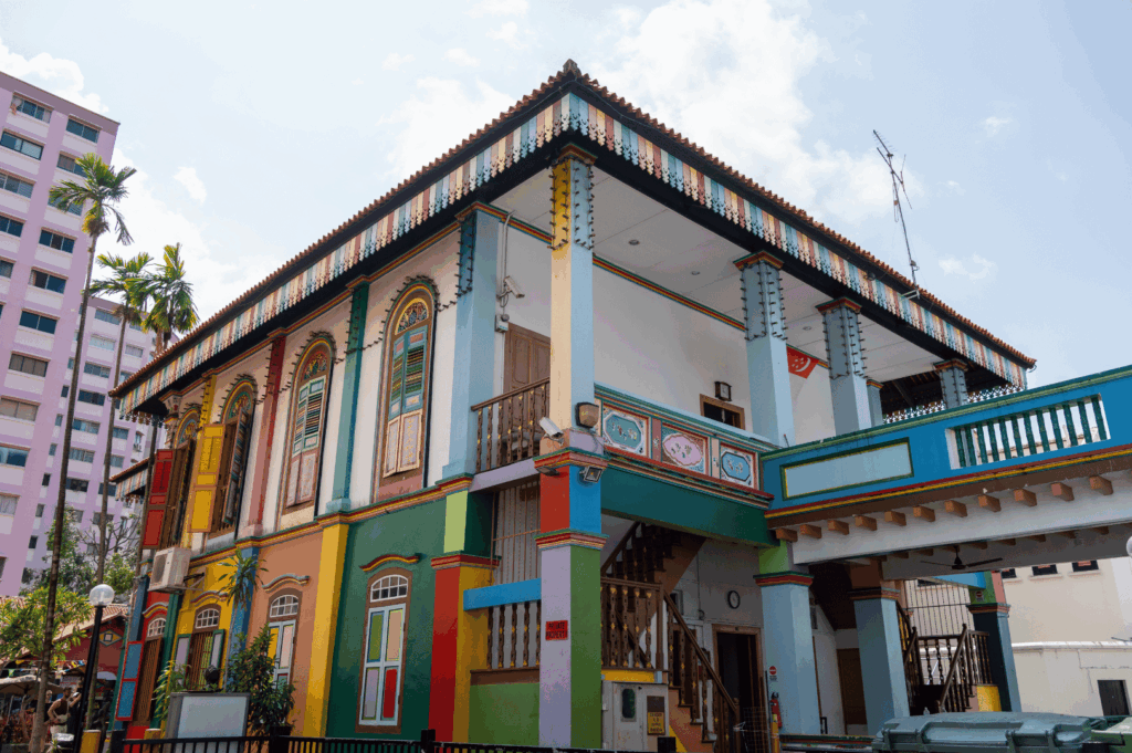 Color-splashed heritage building in Singapore, blending tropical vibrancy with ornate architectural details. Arched windows with multicolored shutters and decorative moldings evoke Peranakan and colonial influences, while the tiled roof and staircase hint at Southeast Asian vernacular design. A visual embodiment of Singapore’s multicultural identity, where tradition and urban dynamism converge.