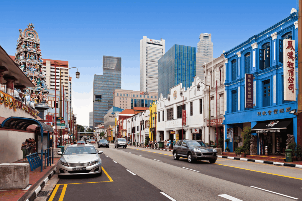Street view of Singapore’s Chinatown, where a vibrant Hindu temple with intricate carvings stands beside colorful shophouses and modern skyscrapers. The juxtaposition of Sri Mariamman Temple’s ornate façade with contemporary high-rises captures the city’s layered cultural landscape. Chinese art centres, heritage businesses, and urban infrastructure converge in a dynamic portrait of Singapore’s pluralistic identity.