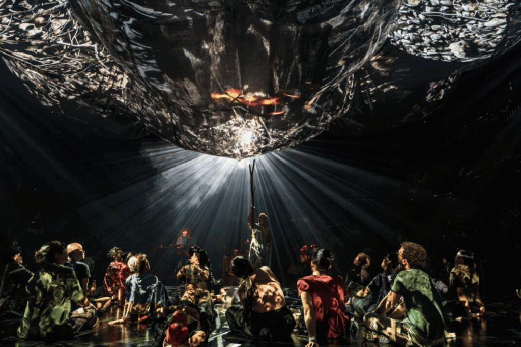 Immersive art installation or theatrical scene in Singapore, where a group sits in a circle beneath a suspended, reflective structure illuminated by dramatic lighting. A central figure reaches upward, bathed in radiant beams that cast surreal shadows across the space. The composition evokes ritual, introspection, and collective experience—blurring the lines between performance, spirituality, and contemporary art.