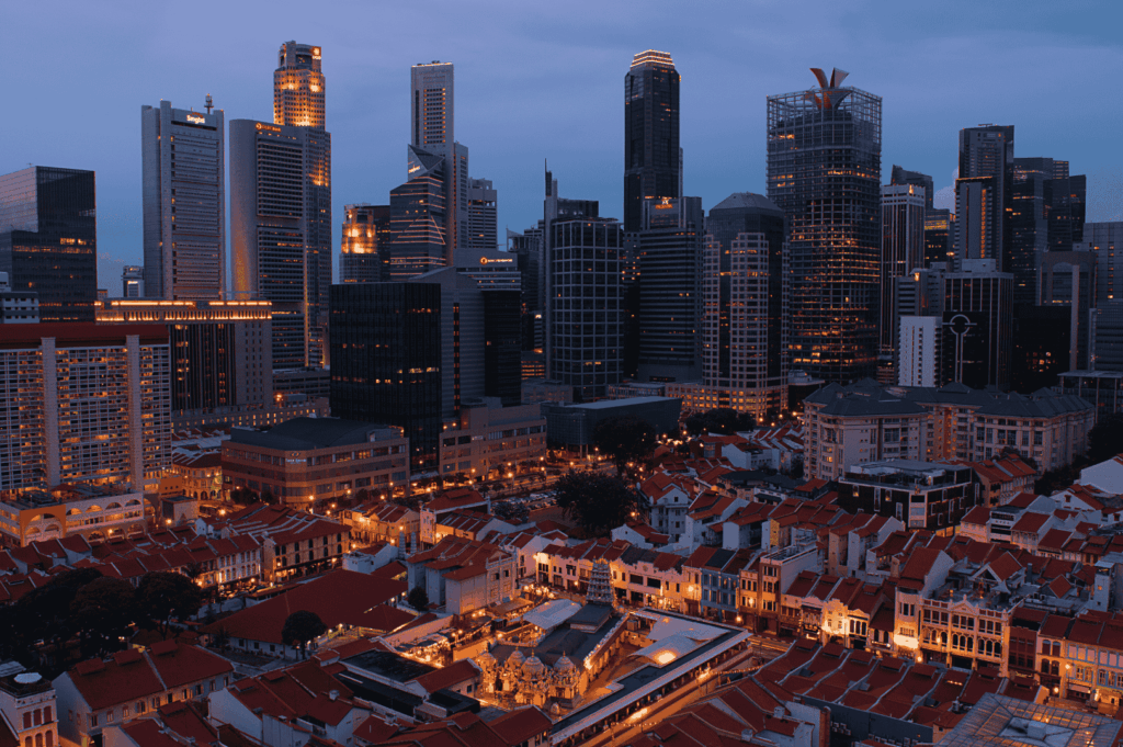 Dusk cityscape of Singapore, where red-roofed heritage shophouses glow in the foreground, arranged in a tight urban grid. Towering modern skyscrapers rise behind them, their illuminated windows signaling a bustling metropolis. The contrast between traditional architecture and contemporary development captures Singapore’s evolving identity—rooted in history, yet reaching toward the future.