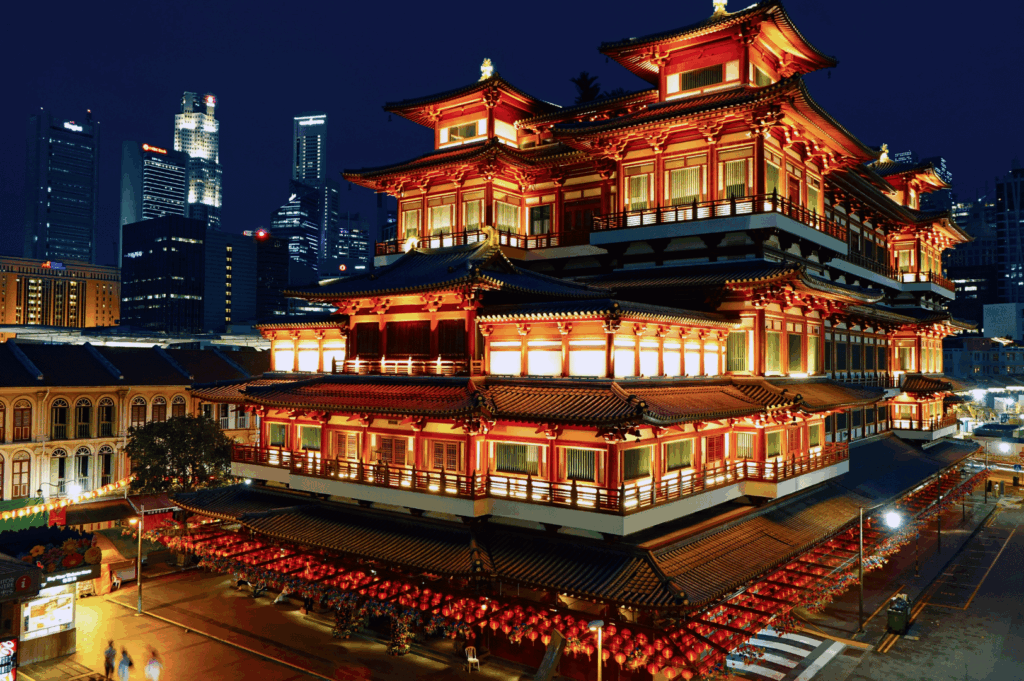 The Buddha Tooth Relic Temple in Singapore’s Chinatown, illuminated in warm golden light against a backdrop of modern skyscrapers. Its multi-tiered rooflines, red columns, and ornate eaves showcase traditional Chinese architectural grandeur. Red lanterns line the street below, infusing the scene with festive energy and spiritual reverence—a striking juxtaposition of heritage and contemporary cityscape.