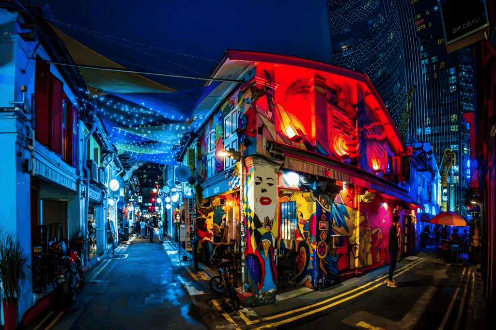 Colourful nightlife street scene in Singapore located in the melting pot of Singapore neighbourhoods such as Chinatown, Kampong Gelam, Little India, Joo Chiat, Tiong Bahru, Holland Village, Robertson Quay, Clarke Quay, Orchard Road, Marina Bay, and the financial district. The bustling area is filled with colourful shophouses, striking murals, hawker stalls, numerous restaurants, bars, and good food, easily accessible and easily accessed with easy access from Changi Airport, and within walking distance to malls, shopping streets, and cultural attractions. Visitors can explore local Peranakan culture, fresh produce markets, and residential area houses in Bukit Timah, Bukit Batok, Bukit Panjang, Pasir Ris, and Paya Lebar, or head south to Sentosa Island and Sentosa Cove hotels, north to nature lovers’ trails, west to Dempsey Hill dining, and east to East Coast Park, Eastpoint Mall, and island escapes. Highlights include the Singapore River, Singapore Zoo, nightlife, and neighbourhoods popular with young couples, expats, and residents. This guide to the country’s towns invites travellers to walk, visit, explore, and indulge in the best neighbourhoods filled with culture, history, and world-class attractions located across the city, north, south, east, and west.