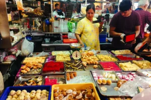 Wide eye‑level shot of a bustling Indian sweets stall in Little India, Singapore, displaying trays of colorful mithai such as laddoos, burfi, jalebi, and milk sweets, while vendors serve customers in a lively market setting.