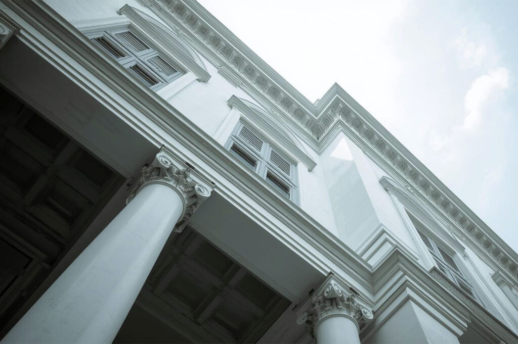 A low-angle view of the white facade of a colonial building, with prominent classical columns and shuttered windows against a bright sky.