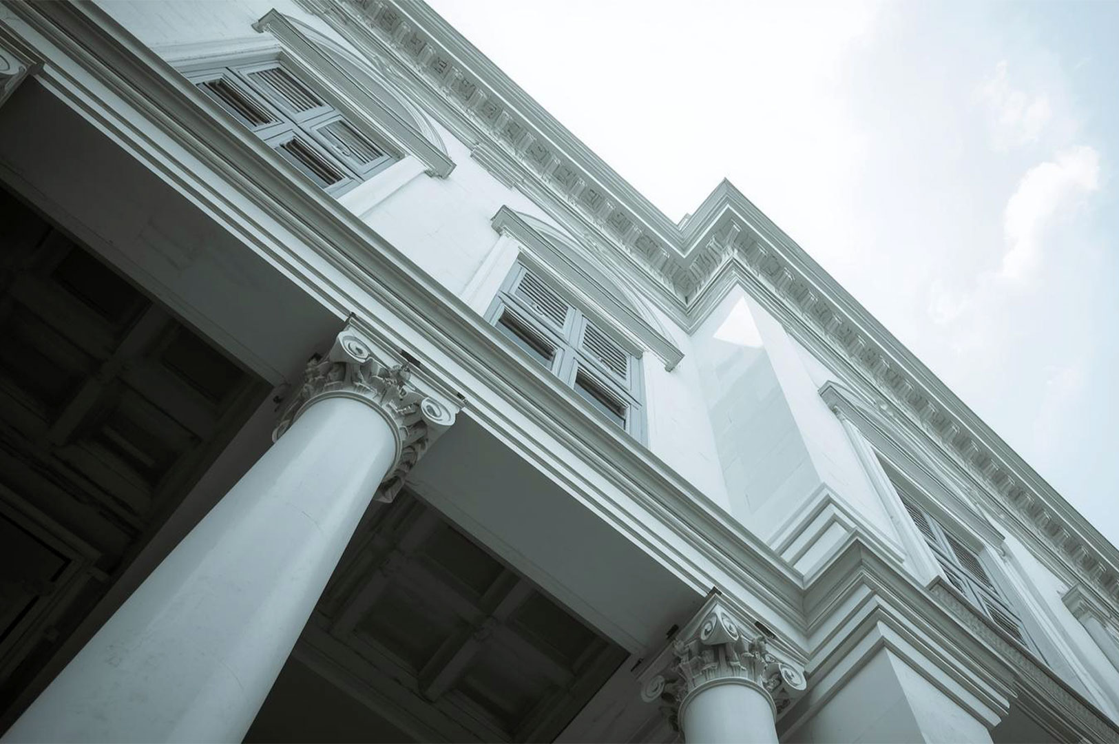 A low-angle view of the white facade of a colonial building, with prominent classical columns and shuttered windows against a bright sky.
