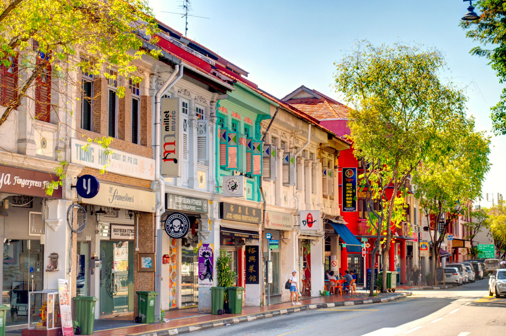 A sunny street scene on Joo Chiat Road in Singapore, lined with vibrant and colourful restored Peranakan shophouses in pastel green, blue, and white, with modern shops on the ground floor.