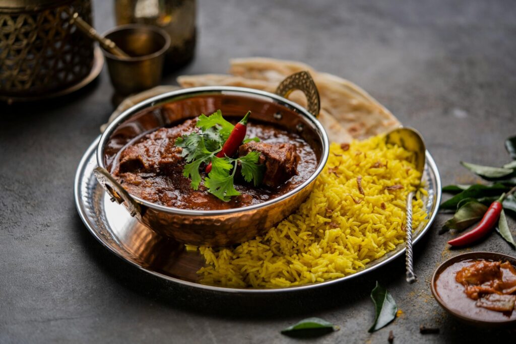 This image features a copper bowl filled with a rich, dark meat curry topped with a fresh red chili and cilantro, served alongside a mound of vibrant yellow saffron rice on a silver platter. Accompanied by a piece of flatbread and a small side dish, the meal is presented on a dark, textured surface with decorative brass elements in the background.