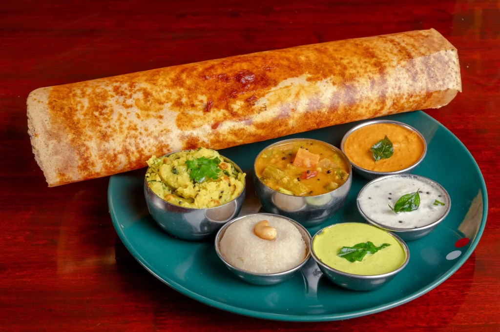Top‑down slightly angled shot of a golden crispy dosa served with assorted chutneys and sambar in small bowls, presented on a green plate, showcasing a classic South Indian breakfast or lunch dish.