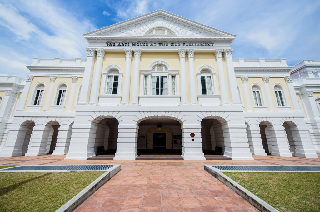 A wide, low-angle shot captures the front facade of The Arts House at The Old Parliament in Singapore under a bright, partly cloudy sky. The building exhibits elegant colonial architecture with a cream-colored main body, white trim, and a prominent pediment atop a series of columns. Large arched walkways form the ground level entrance, leading to a darker interior. In the foreground, a terracotta-tiled pathway is bordered by neatly trimmed green grass, leading directly to the building's entrance. The name "THE ARTS HOUSE AT THE OLD PARLIAMENT" is visible above the central section of the building.