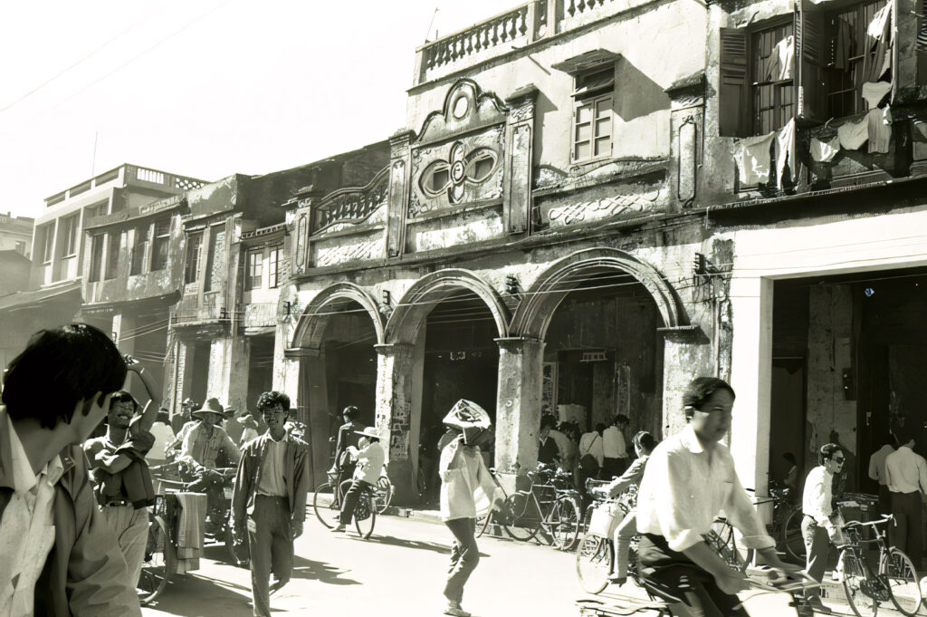 Black and white historical photo of a busy street in Hainan (or early Singapore/Malaysia), showing traditional shophouse architecture with arched walkways and people on bicycles.