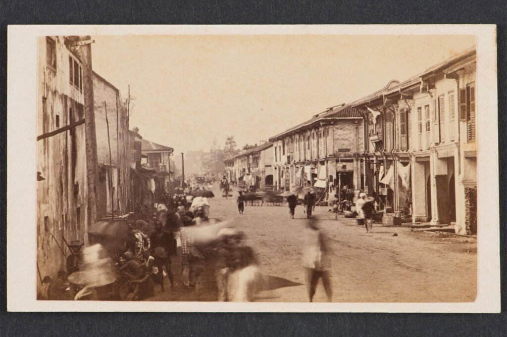 A vintage, sepia-toned photograph of early shophouses on South Bridge Road, Singapore, showing a bustling, unpaved street with pedestrians and traditional two-story buildings from the 19th century