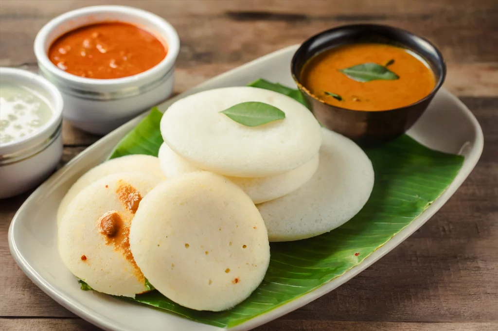 Close-up eye‑level shot of soft steamed idli arranged on a banana leaf, served with sambar and coconut chutney, emphasizing comfort food and authentic South Indian flavors.