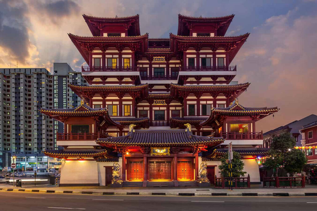The magnificent, multi-tiered red and gold architecture of the Buddha Tooth Relic Temple in Singapore's Chinatown, set against a sunset sky and surrounding modern high-rises.
