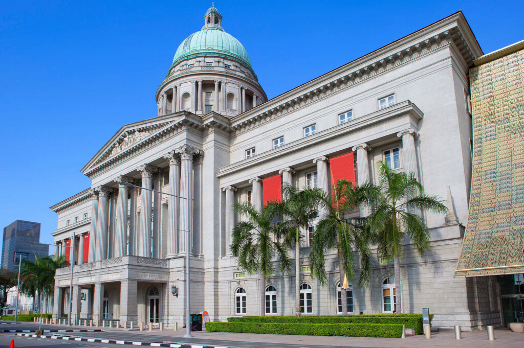 The former Supreme Court building in Singapore, a grand neoclassical structure, is viewed from a low angle under a clear blue sky. The prominent building features a large green dome, multiple columns, and white stone facade. Bright red banners hang vertically from some of the windows, adding a pop of color to the otherwise stately architecture. Lush green palm trees line the front of the building, and a paved road with a striped curb is visible in the foreground.