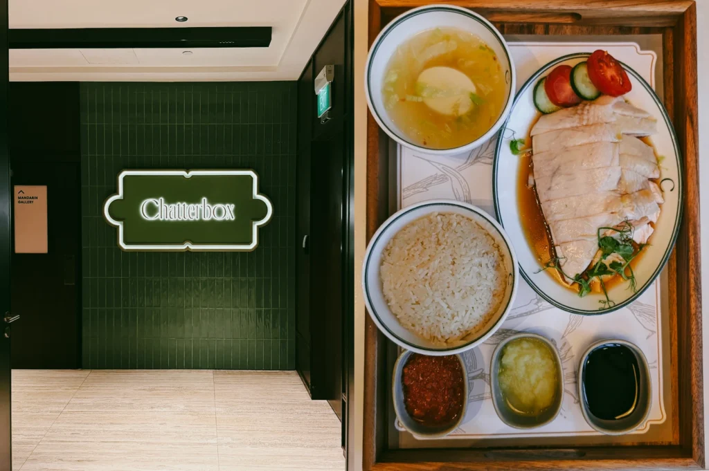 Left: An eye-level, medium shot of the modern, minimalist entrance to a Chatterbox restaurant, highlighting a green tiled wall with a backlit signature logo sign. Right: A top-down, close-up shot of a complete Hainanese chicken rice set arranged on a wooden tray, including a bowl of rice, a bowl of broth, sliced chicken garnished with cucumber and tomato, and three small dishes containing chili, ginger, and dark soy sauce.”