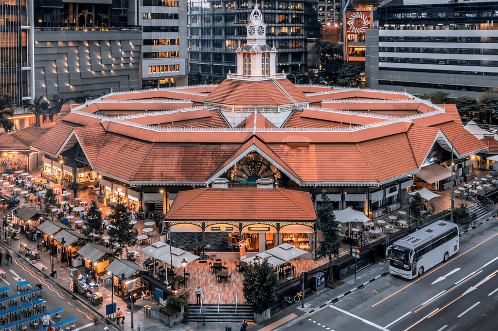 An aerial view captures Lau Pa Sat, a historic hawker center in Singapore, at dusk or early evening, illuminated with warm lights. The distinctive octagonal building with its terracotta-tiled roof and central clock tower stands out amidst modern city buildings in the background. Around the hawker center, numerous outdoor food stalls with seating areas are bustling with activity, extending to the left side of the image. A street with a white tour bus is visible on the right, and the surrounding urban environment blends historic charm with contemporary city life.