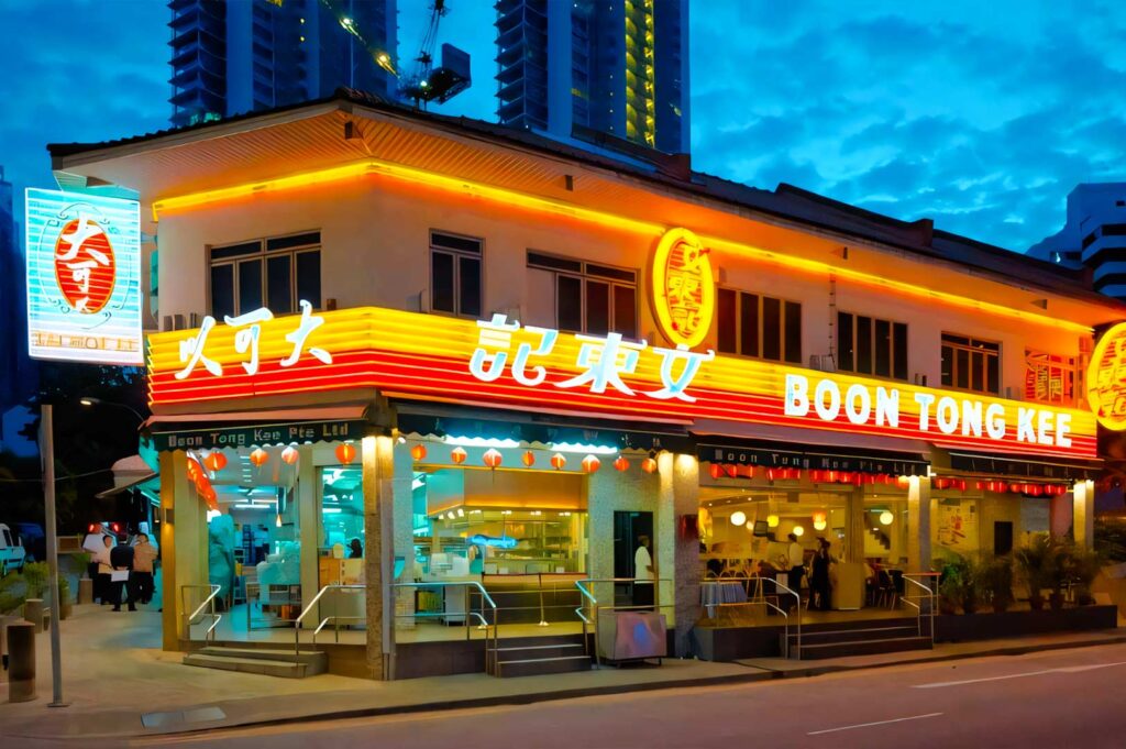Exterior of Boon Tong Kee restaurant at dusk, featuring bright neon signage, illuminated windows, and a vibrant city backdrop, showcasing one of Singapore’s most popular Hainanese chicken rice dining spots.