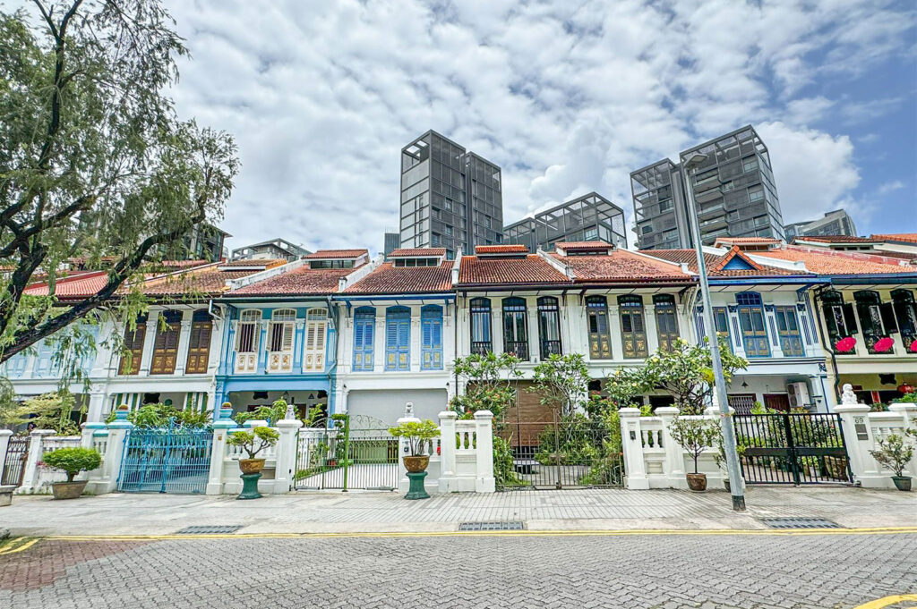 A row of beautifully preserved Peranakan residential shophouses on Emerald Hill, Singapore, showing their colourful facades and gated entrances, with the tall skyscrapers of the modern city skyline rising in the background