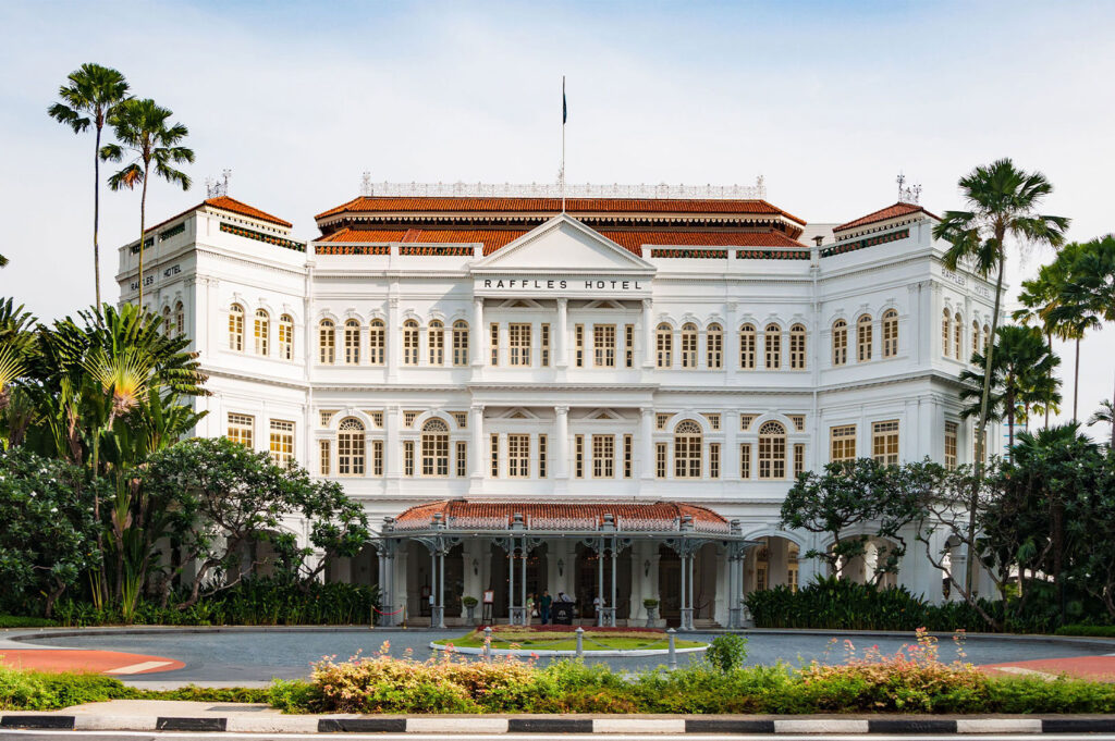 The iconic Raffles Hotel in Singapore is captured in a wide shot, showcasing its grand colonial architecture under a bright sky. The elegant white facade, adorned with numerous windows, is framed by lush tropical greenery, including tall palm trees on either side. The hotel's prominent entrance features a covered driveway and intricate ironwork details. A semicircular garden with well-maintained foliage sits in the foreground, separating the hotel from the paved road.