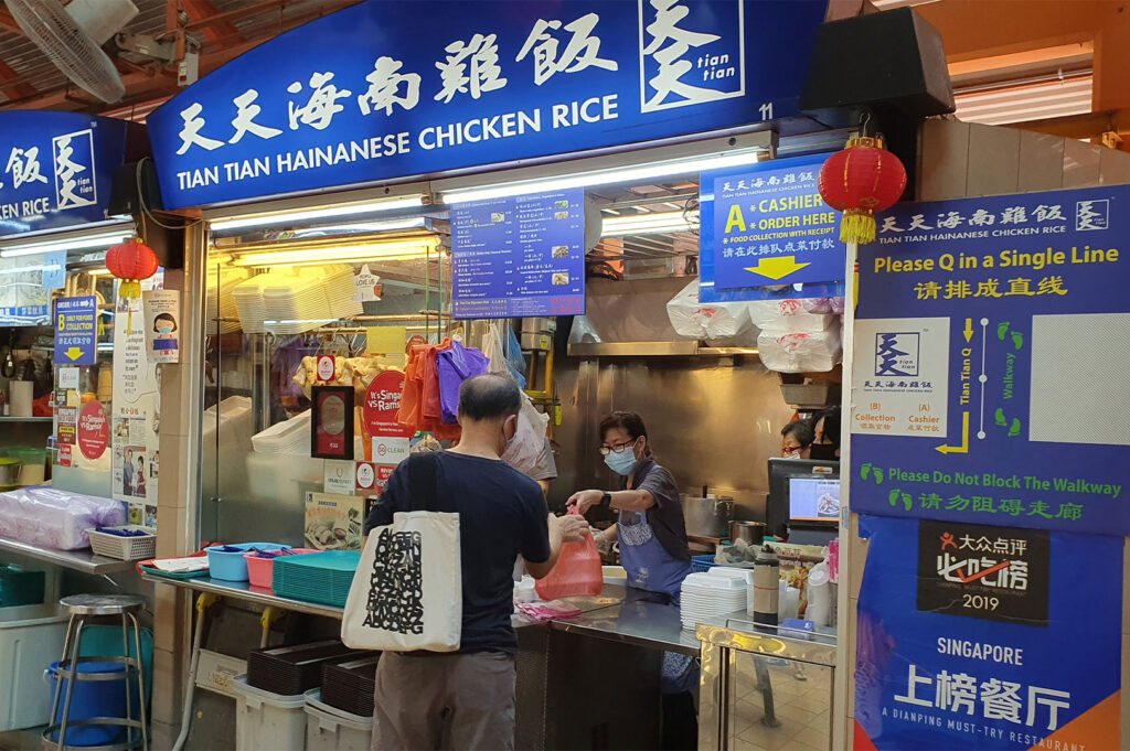 The busy counter of the famous Tian Tian Hainanese Chicken Rice stall in Maxwell Food Centre, showing a cashier and a customer waiting to pick up food.