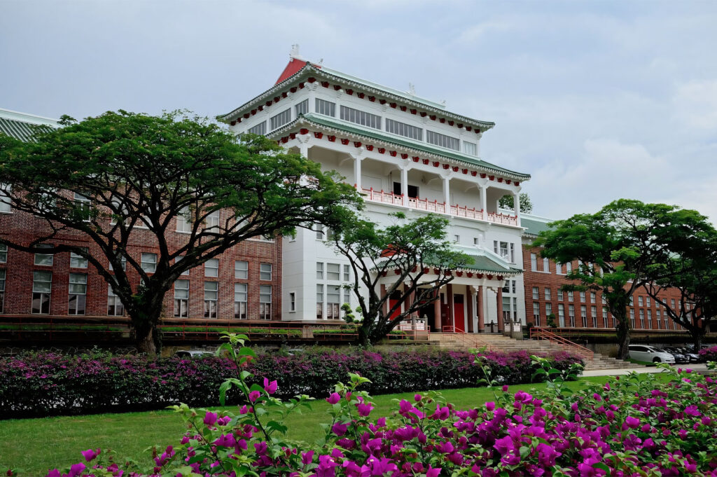 The stately, colonial-style Chinese Heritage Centre building, featuring white walls, a red-tiled roof, and surrounding lush green lawns and bright pink flowers.